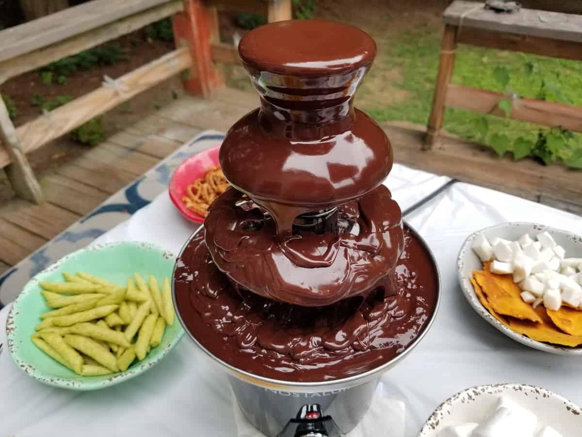 3 tiered chocolate fountain with multiple foods to dip in the chocolate pictured in the background.