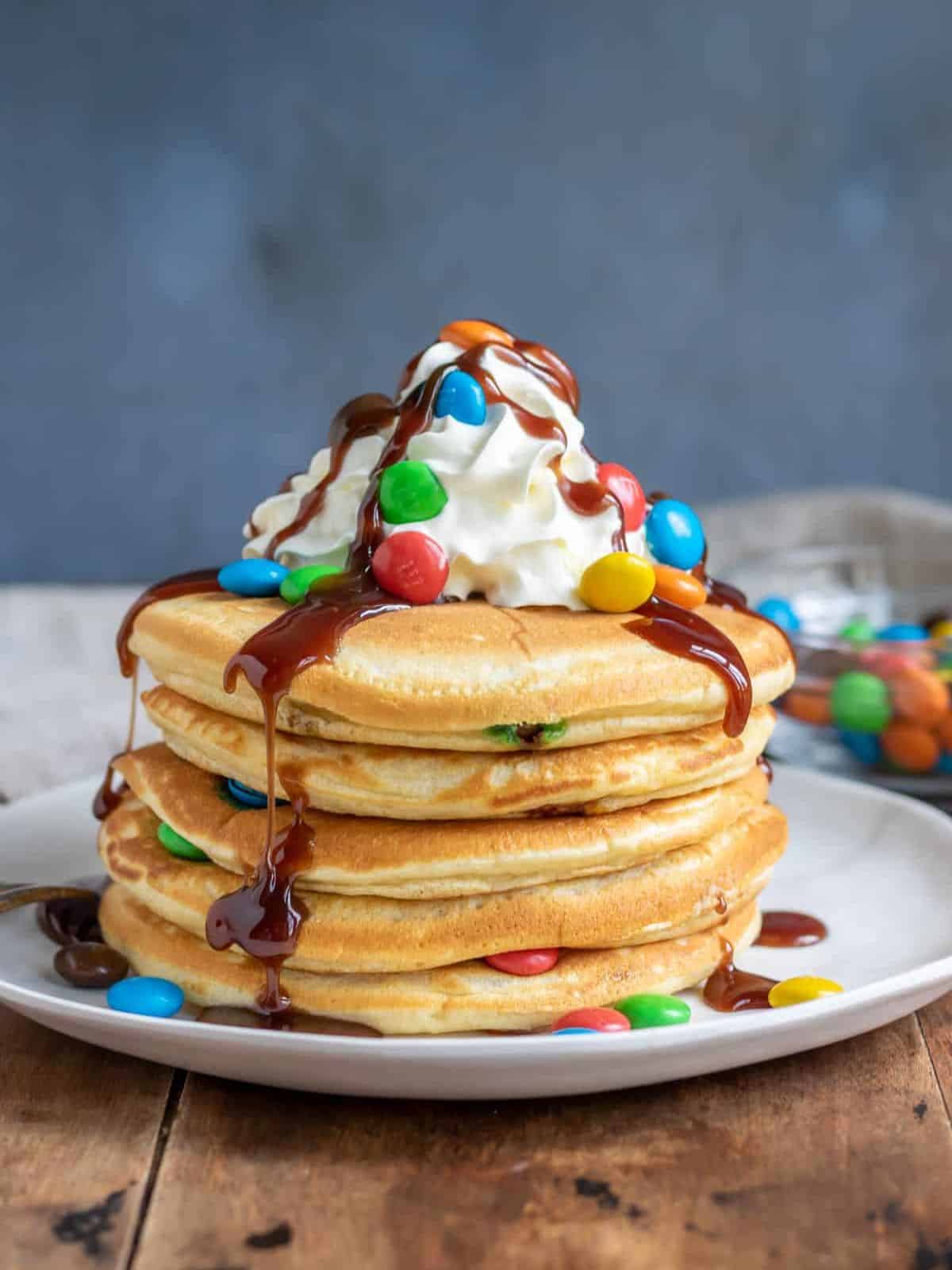 Stacked pancakes topped with maple syrup, M&Ms, and whipped cream.
