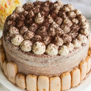 a luscious whole tiramisu cake dessert made with moist vanilla sponge soaked in coffee and a velvety mascarpone filling, with lady fingers on the edge of the cake, topped with cocoa powder.