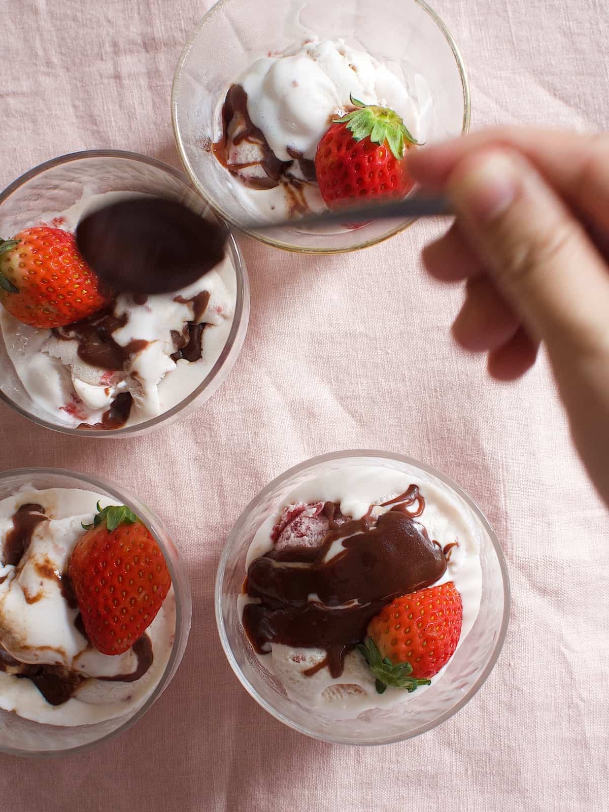 A spoon pouring fudge over four sundaes topped with strawberries.