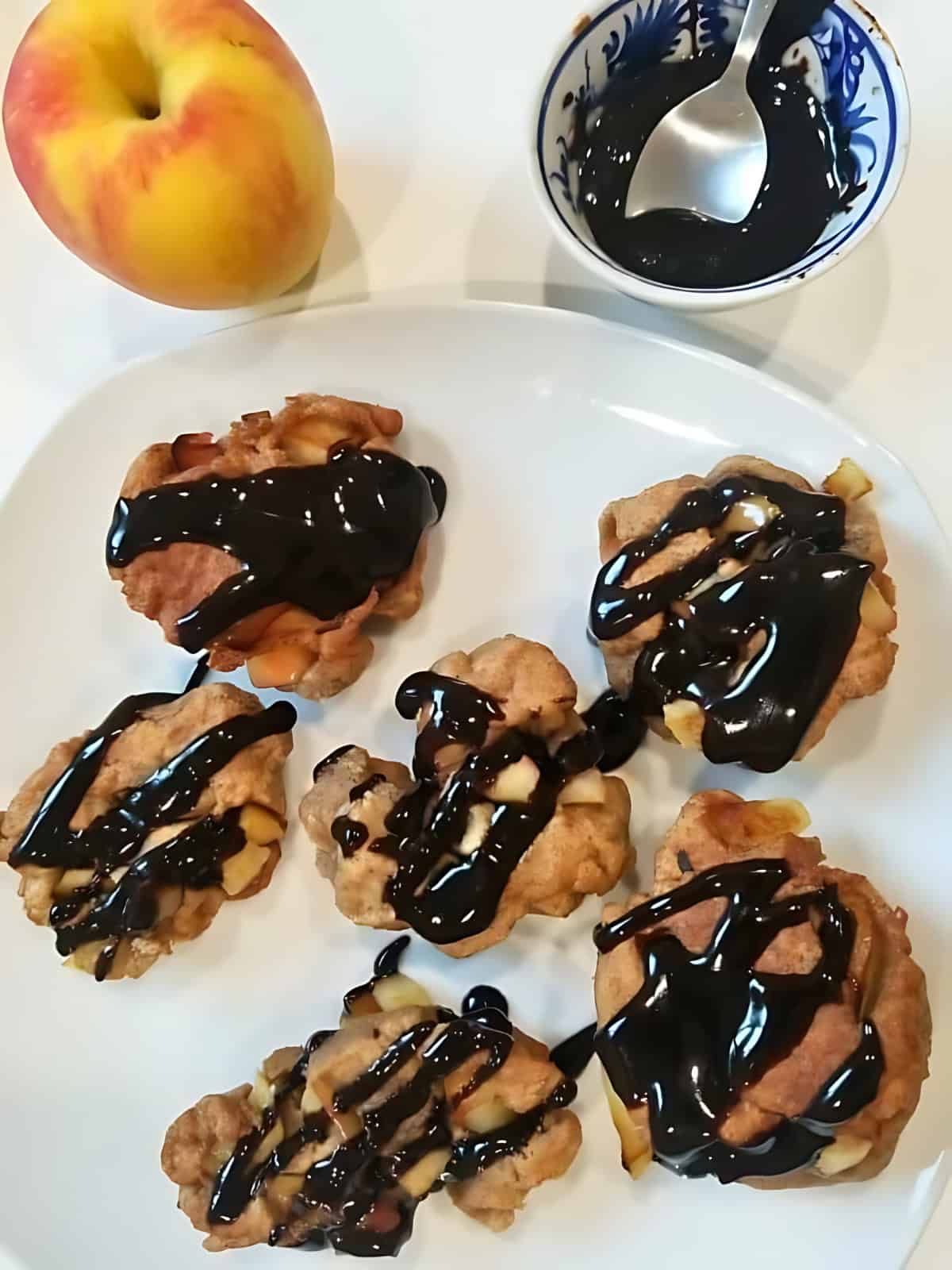Apple fritters drizzled with chocolate fudge on a plate, next to a fresh apple.