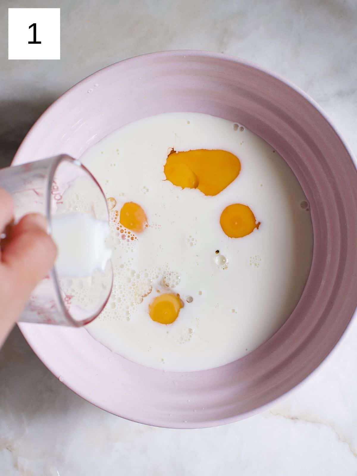 A person pouring milk to a bowl of eggs mixed with maple syrup.