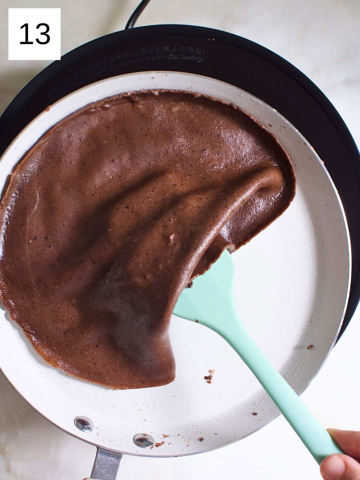 A person flipping a chocolate crap with a spatula in a heated pan.