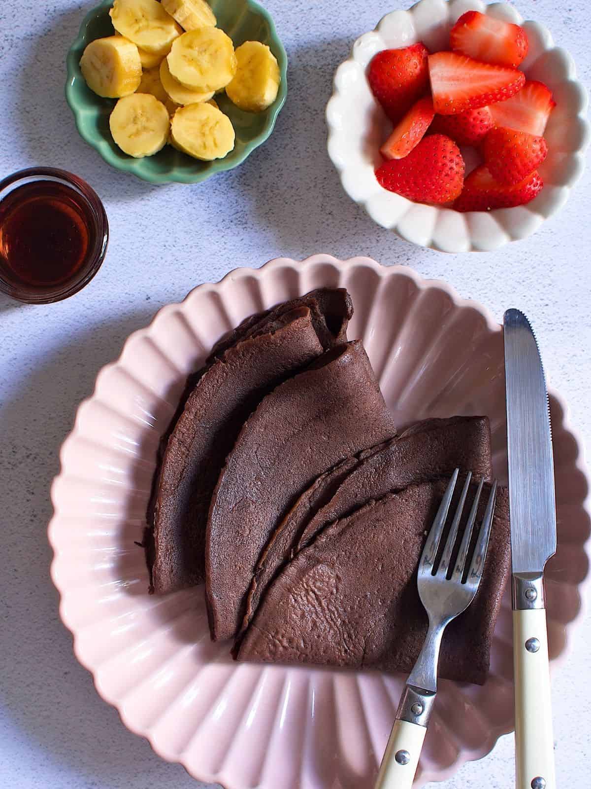 Folded chocolate crepes on a plate next to a bowl of sliced bananas, strawberries, and maple syrup.