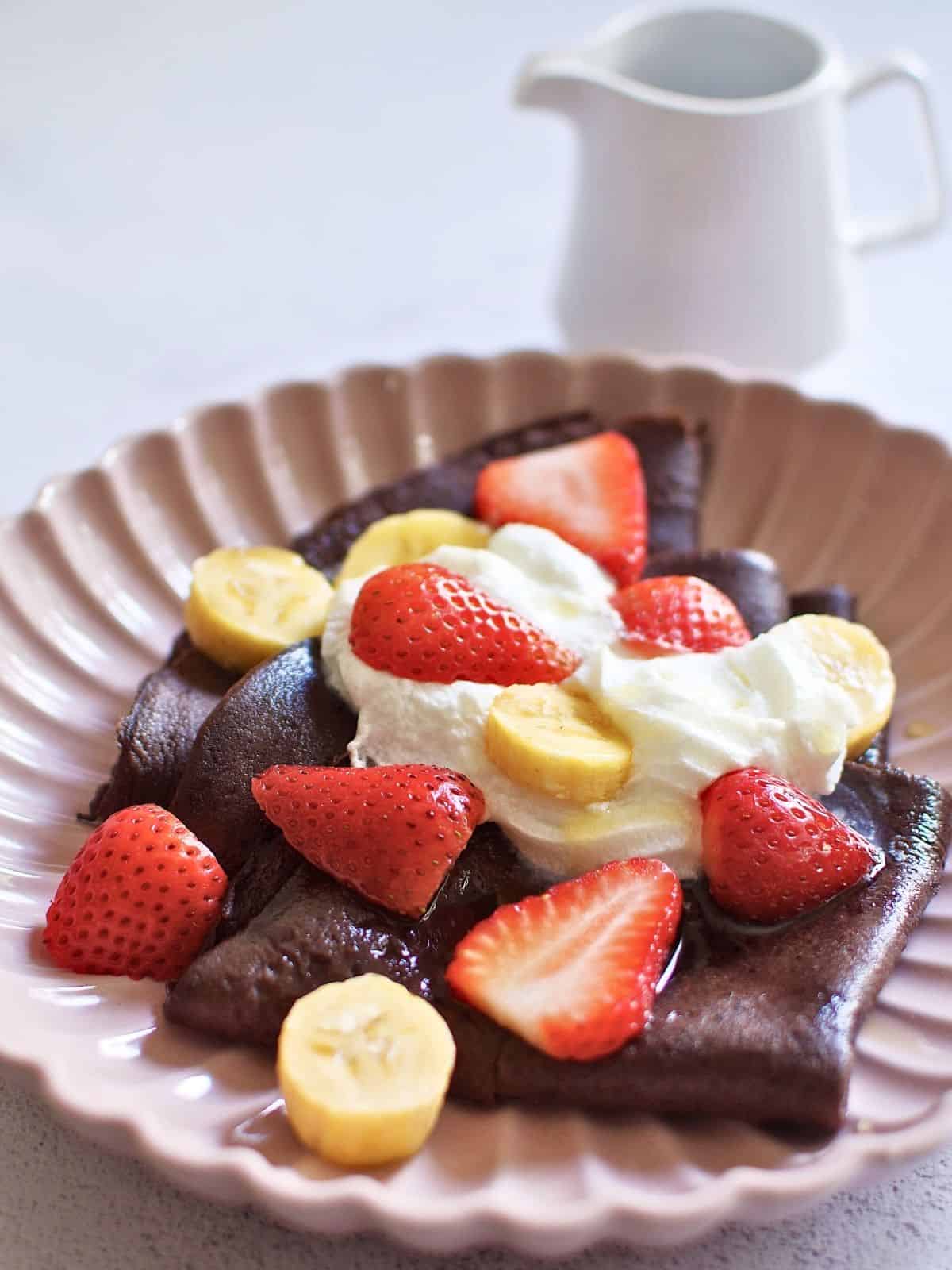 Chocolate crepes topped with sliced banana and strawberries on a plate next to maple syrup.