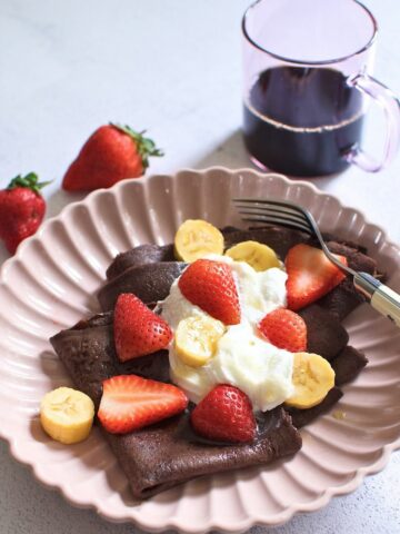 Chocolate crepe topped with topped with sliced bananas and strawberries on a plate.