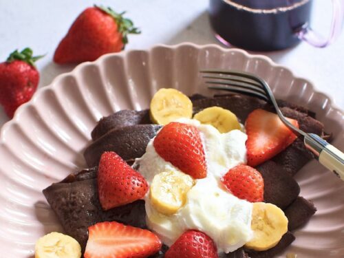 Chocolate crepe topped with topped with sliced bananas and strawberries on a plate.