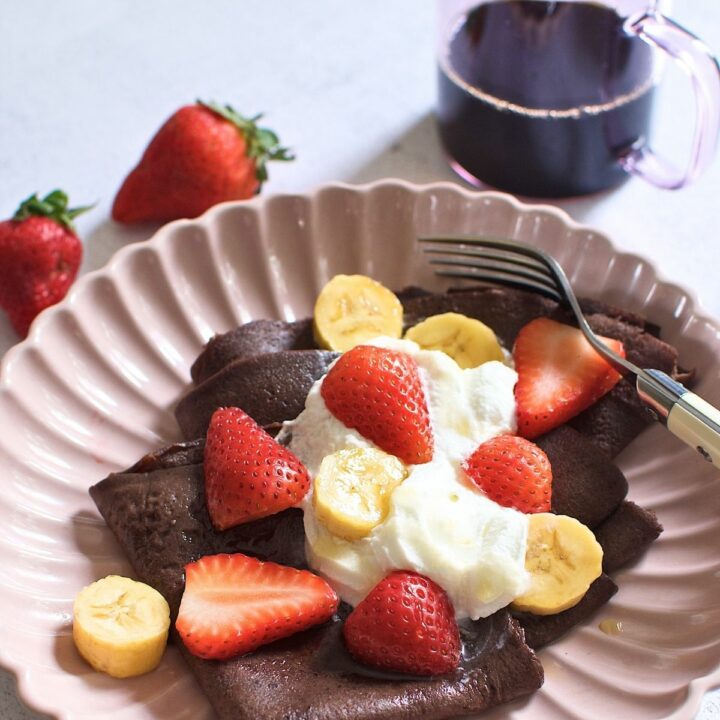 Chocolate crepe topped with topped with sliced bananas and strawberries on a plate.