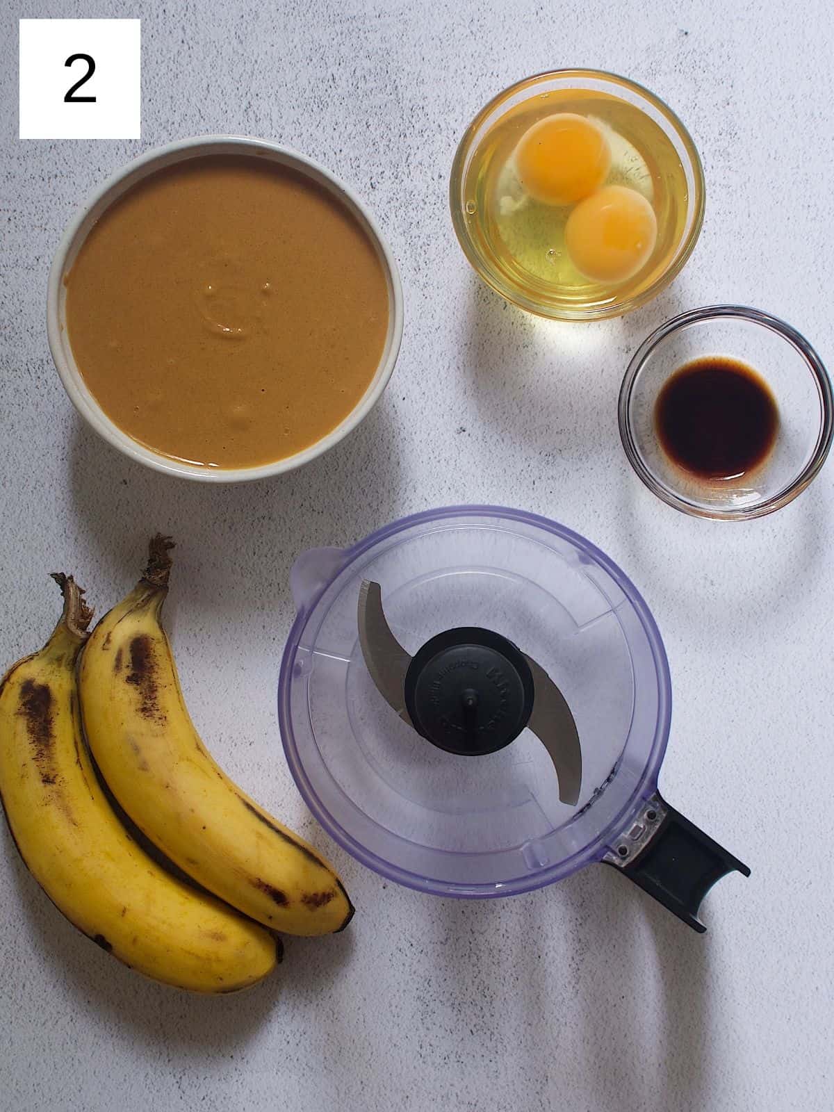 A pair of bananas next to a bowl of nut butter, eggs, vanilla, and a food processor.