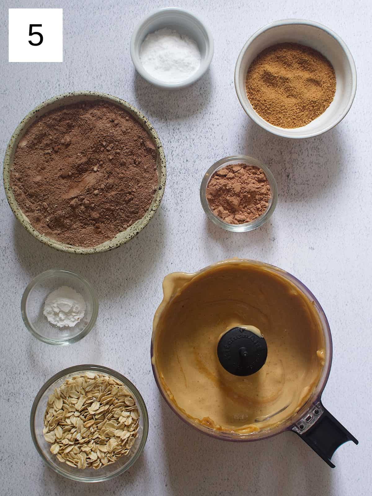 Banana muffin batter next to a bowl of chocolate protein powder, baking powder, baking soda, cocoa sugar, cacao powder, and whole rolled oats.