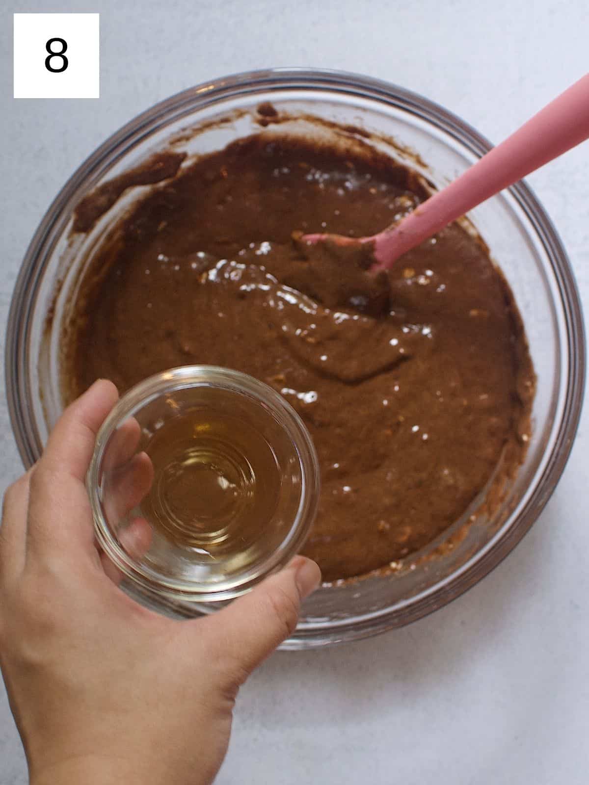 A person about to pour a small bowl of apple cider vinegar into a bowl of banana chocolate muffin batter.