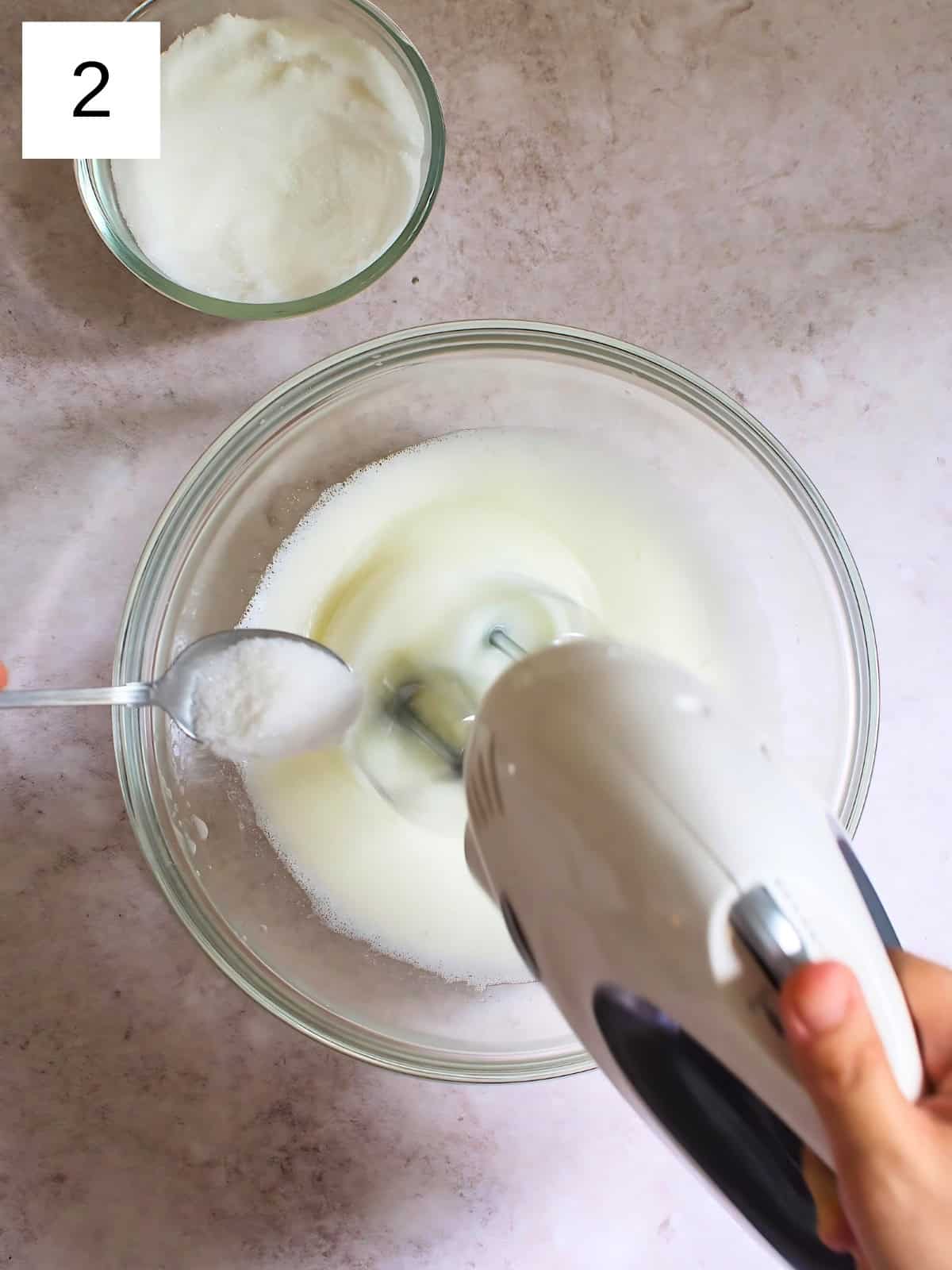 A person beating a bowl of egg whites using a hand mixer while gradually adding a spoon of sugar.