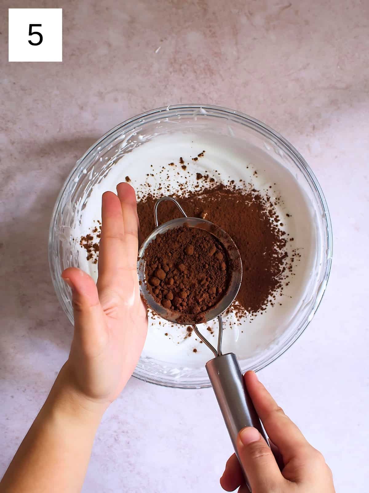 A person sprinkling cocoa powder with a sift on top of a bowl of meringue.