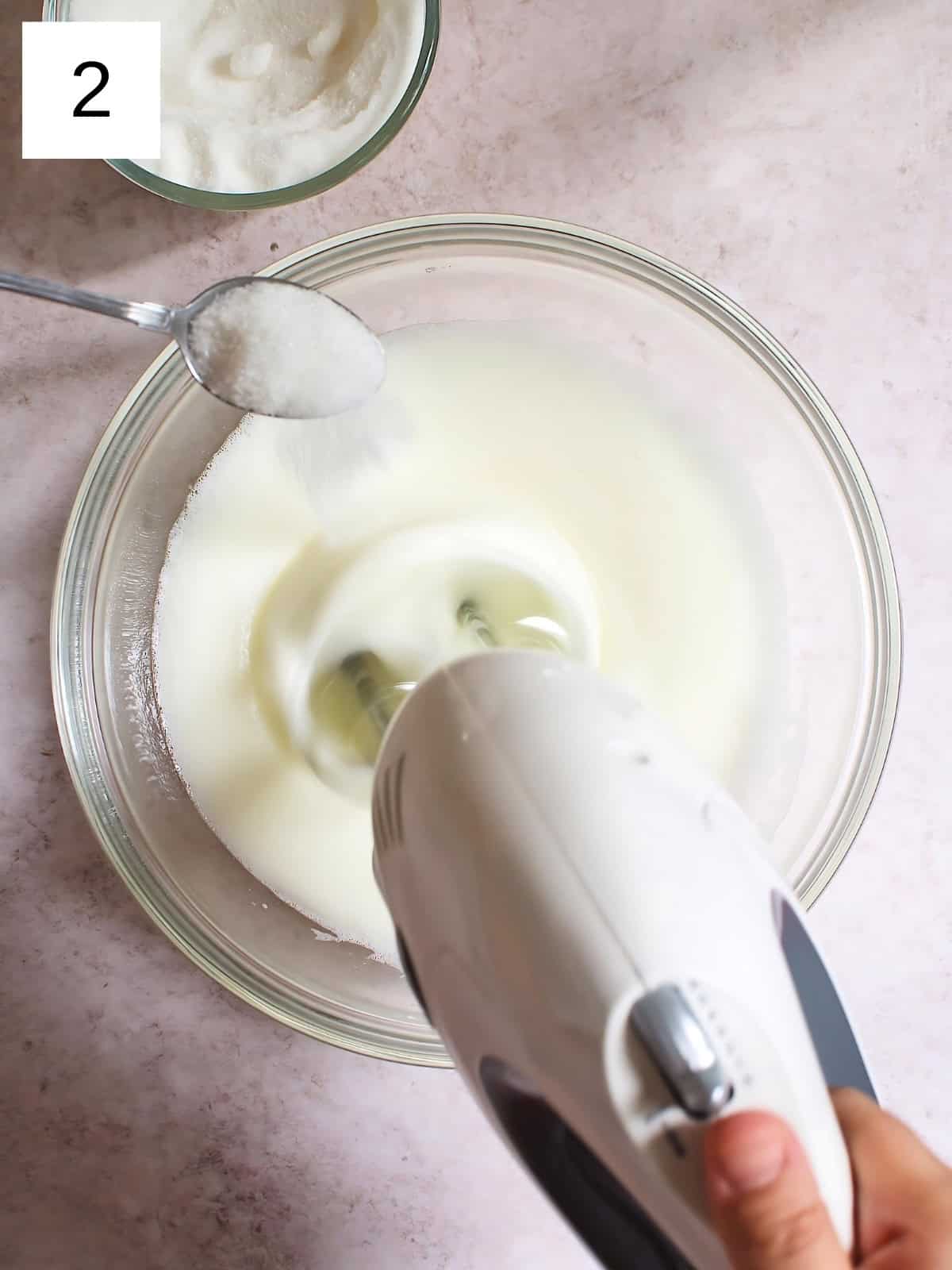 A person whisking egg whites in a bowl while gradually adding a spoon of sugar.