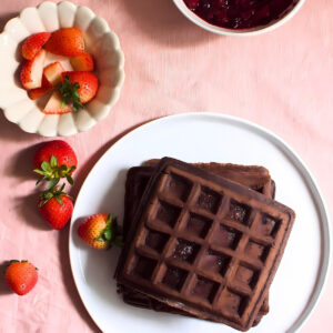 Stacked chocolate waffles in a plate next to strawberries and a bowl of jam.