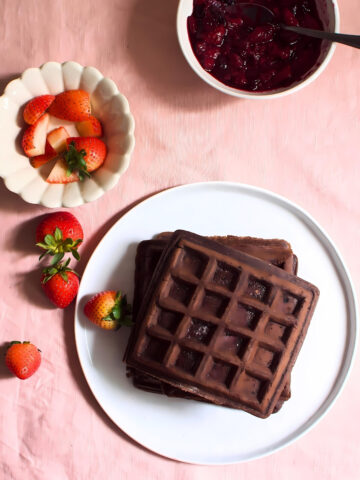 Stacked chocolate waffles in a plate next to strawberries and a bowl of jam.