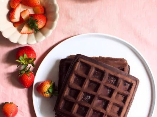 Stacked chocolate waffles in a plate next to strawberries and a bowl of jam.