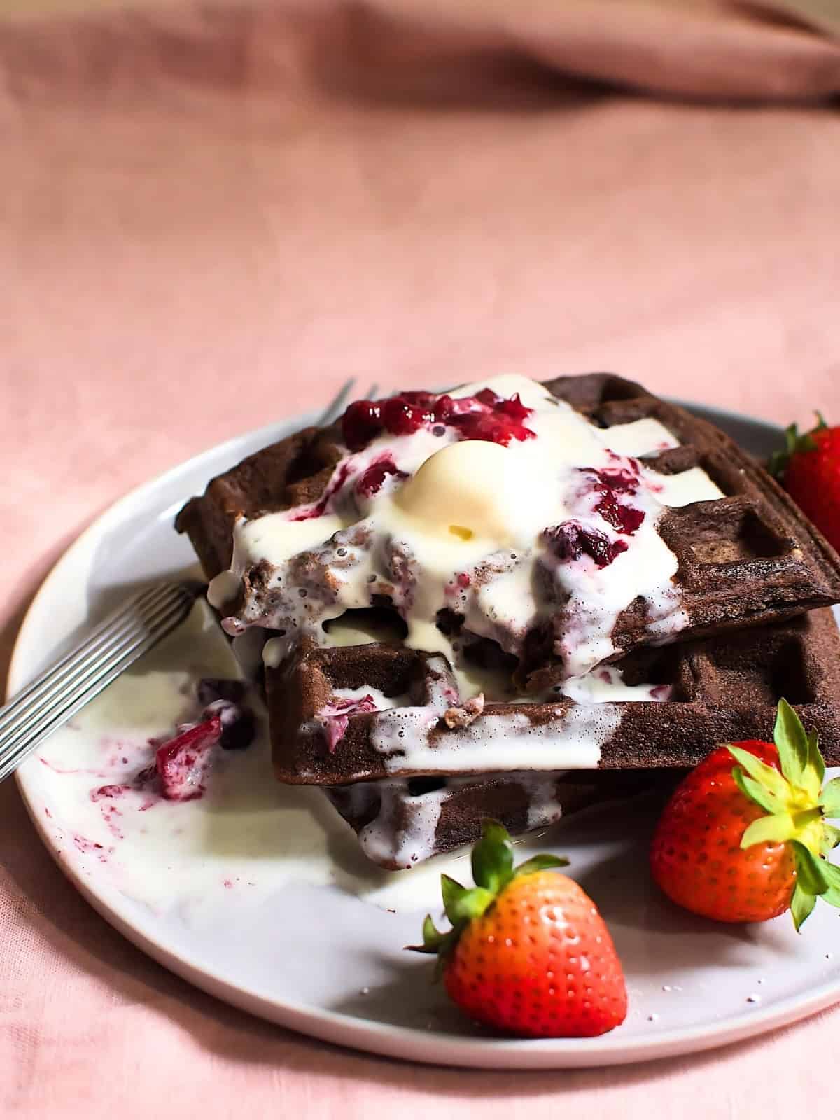 Stacked chocolate waffles in a plate topped with butter cream and jam next to strawberries.