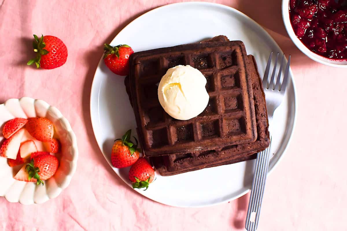 Stacked chocolate waffles in a plate topped with butter cream next to strawberries.