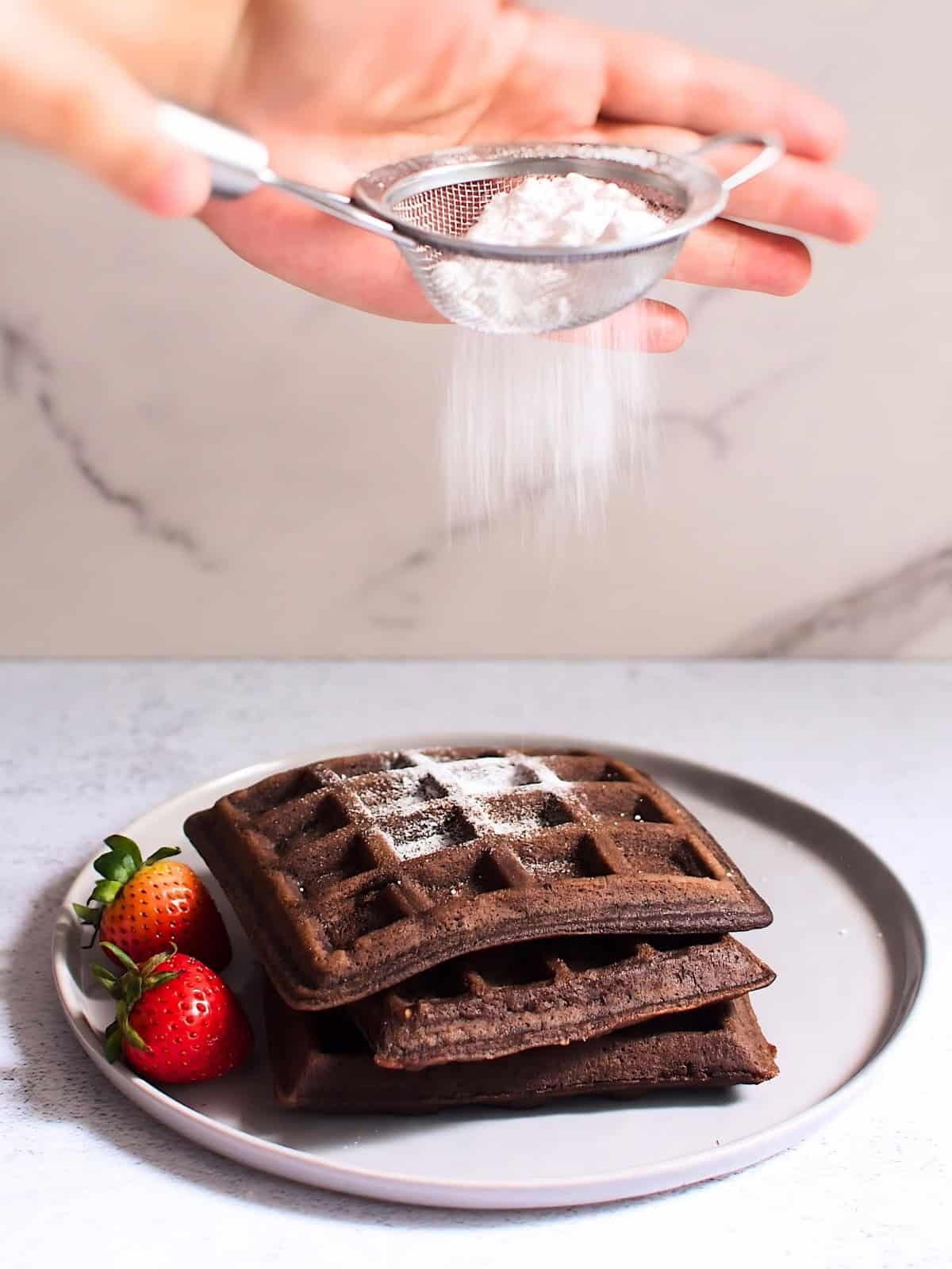 Chocolate waffles in a plate with strawberries sprinkled with powdered sugar.