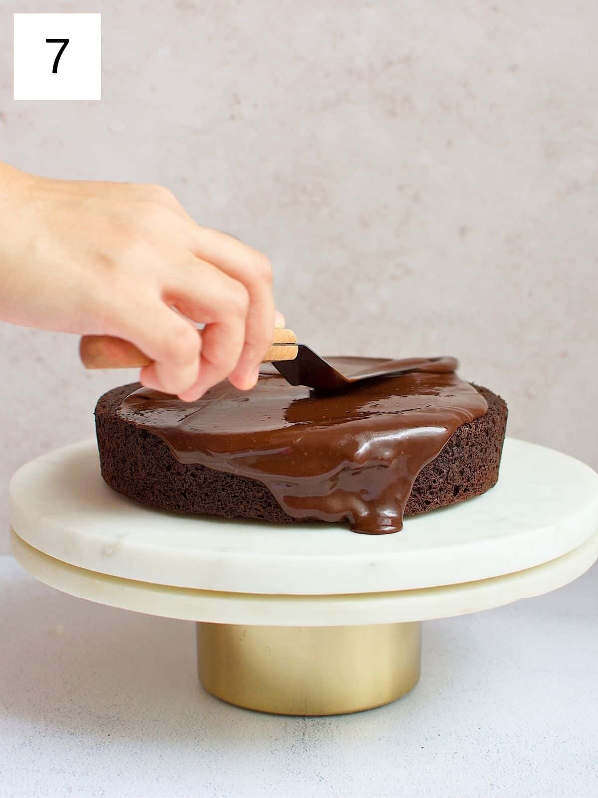 A person spreading chocolate ganache evenly on a chocolate cake.