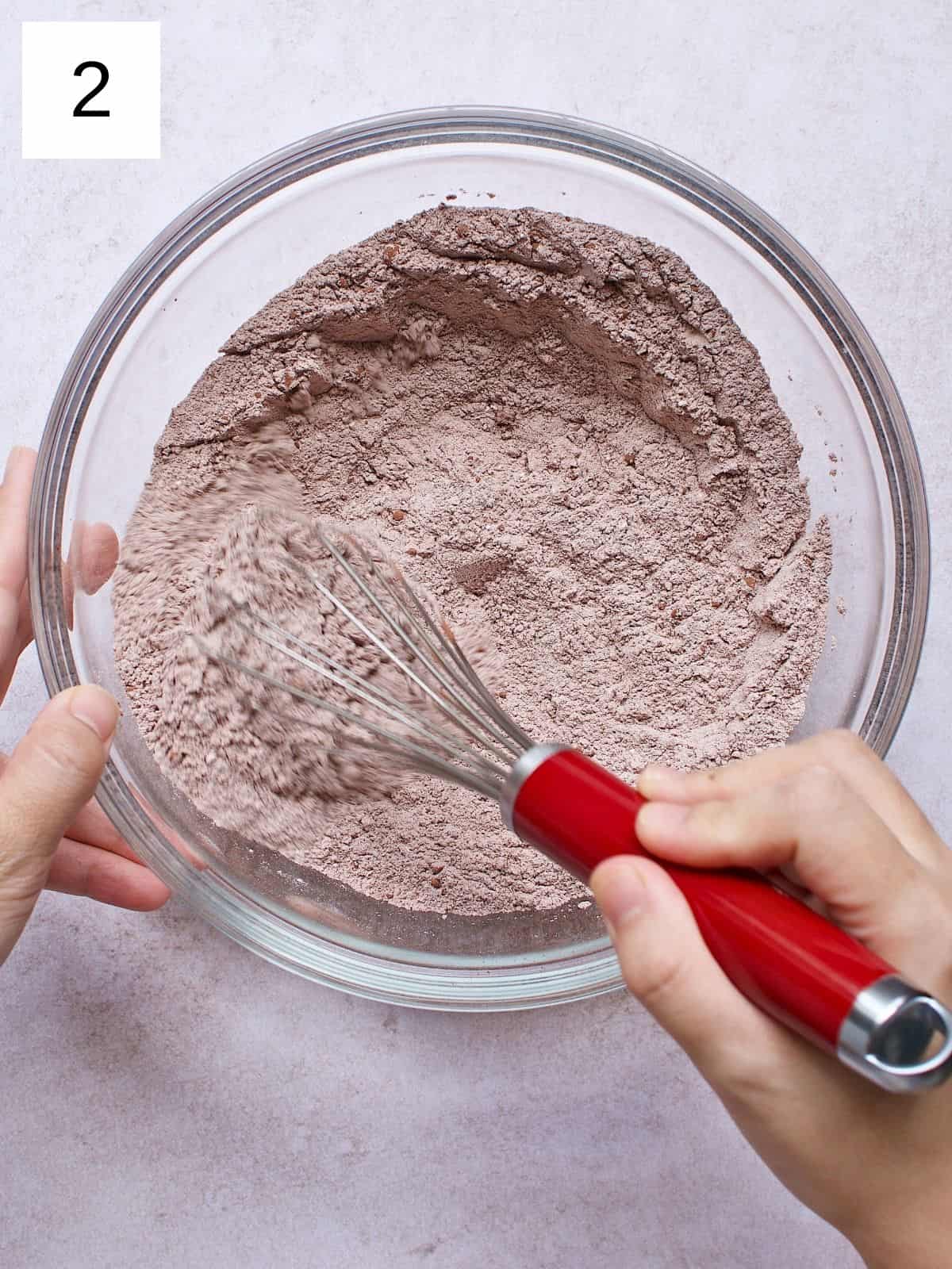 A person whisking flour, cocoa powder, sugar, baking powder, salt, and baking soda in a bowl.