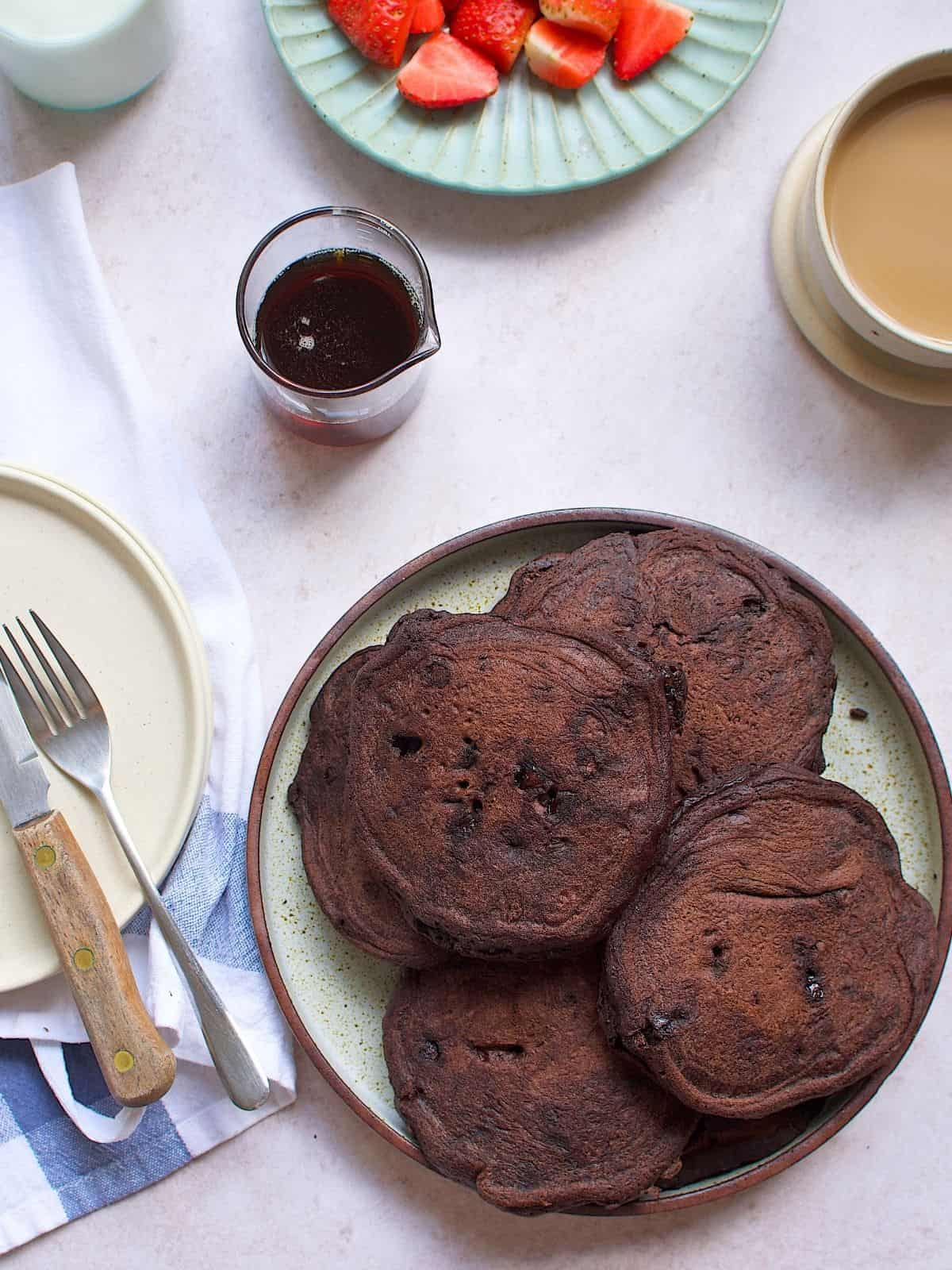 Stacks of chocolate pancakes on a plate next to maple syrup.