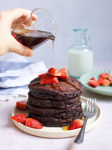 A person pouring maple syrups over a stack of chocolate pancakes with strawberries.