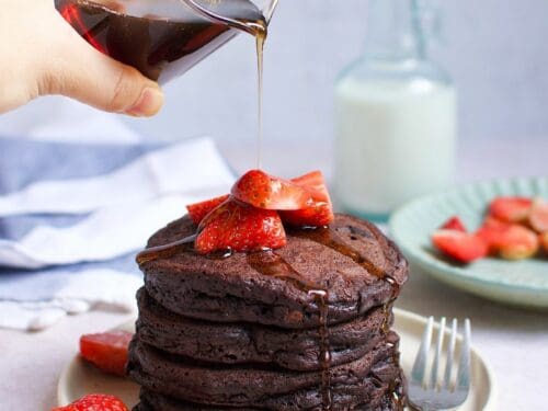 A person pouring maple syrups over a stack of chocolate pancakes with strawberries.