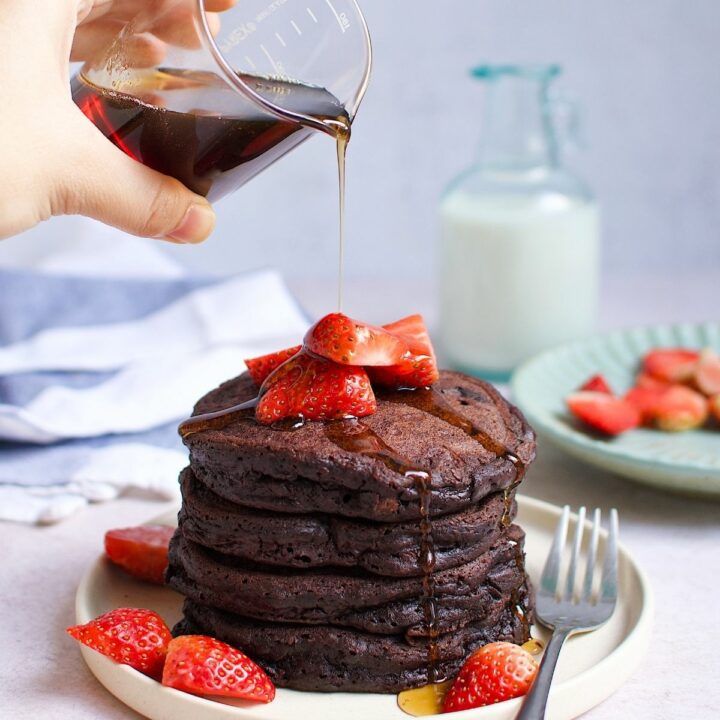 A person pouring maple syrups over a stack of chocolate pancakes with strawberries.