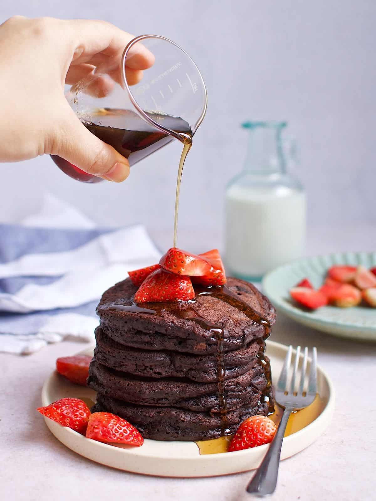 A person pouring maple over stacked chocolate pancaked wtih strawerries on a plate.