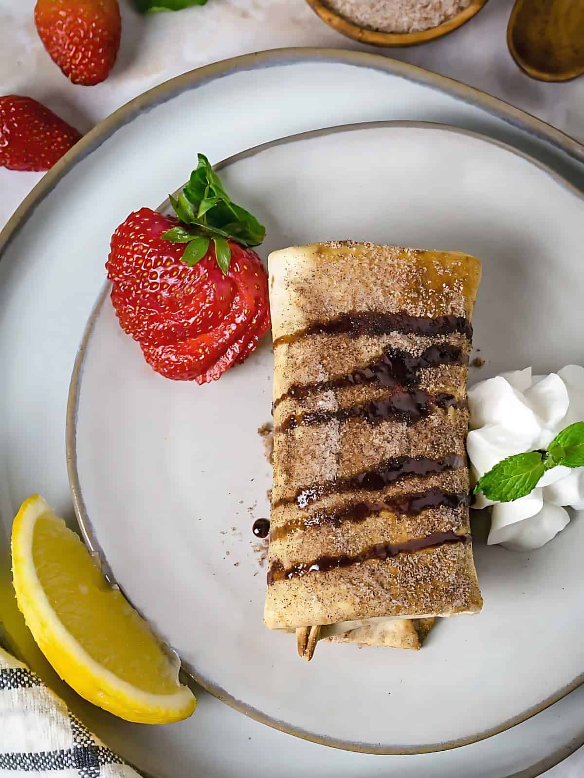 Strawberry cheesecake chimichangas drizzled with chocolate syrup on a plate next to strawberries.