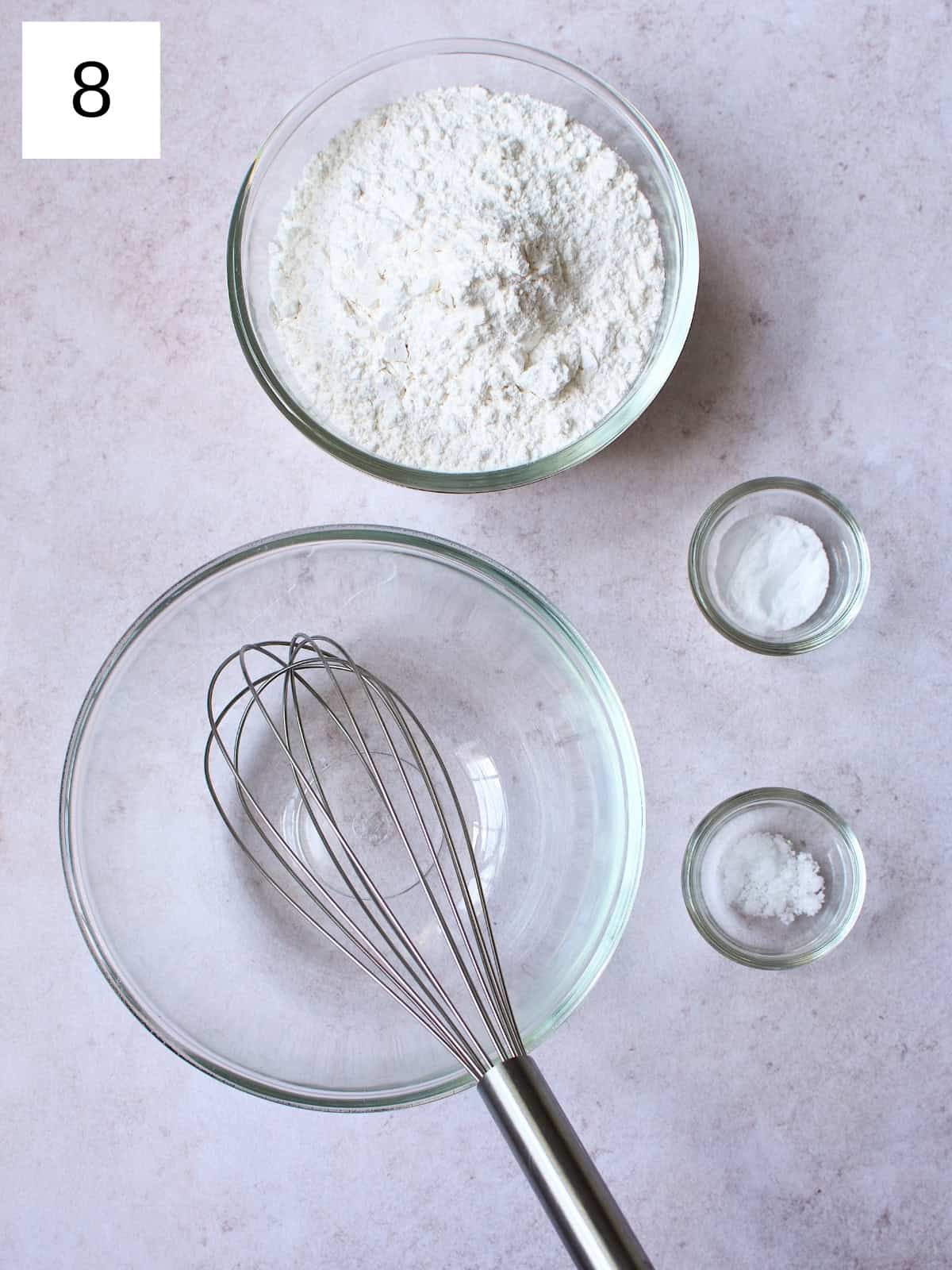 Two bowls, one empty and one filled with all-purpose flour, alongside two small saucers holding baking soda and salt, arranged on a clean surface, ready for baking preparation.