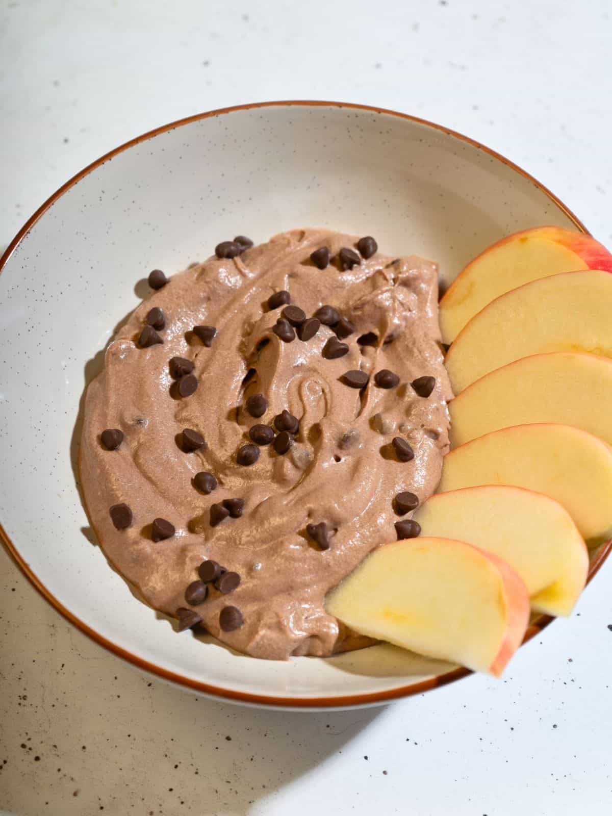 Creamy brownie batter dip with chocolate chips in a bowl, served with fresh apple slices on the side.