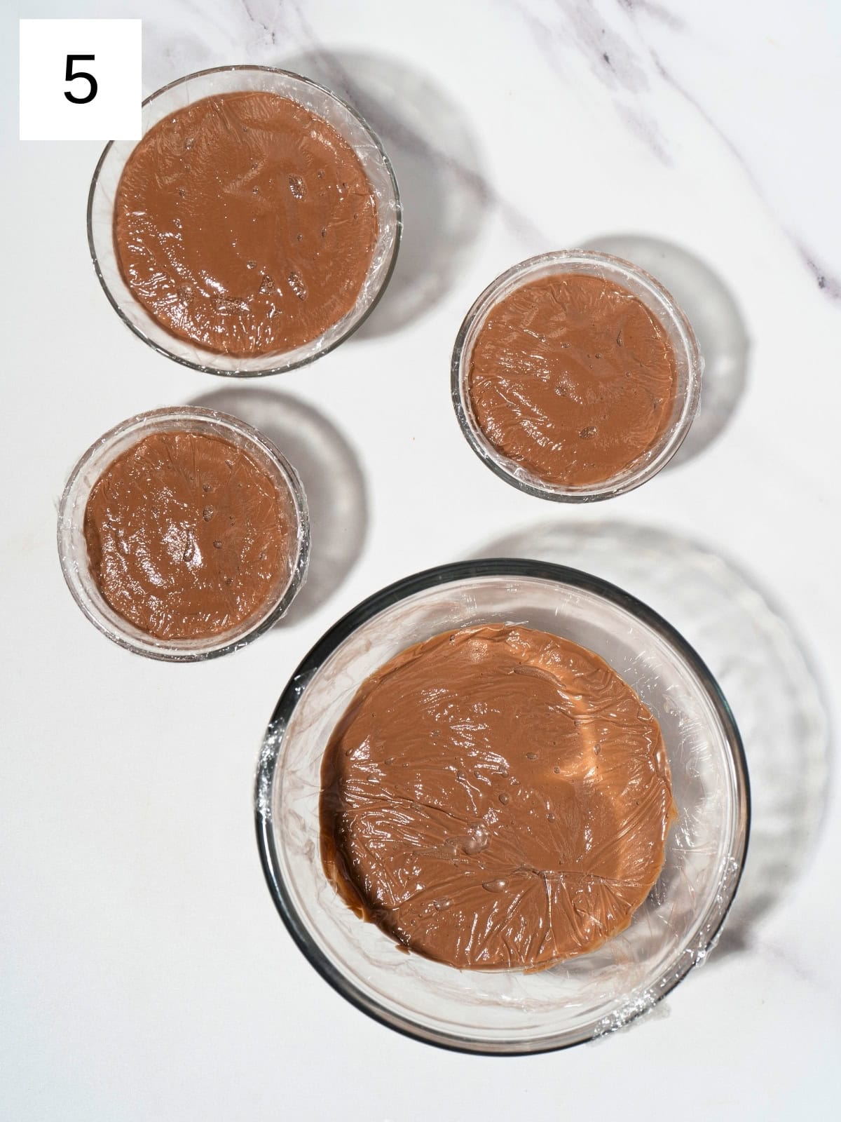 Pudding mixture in four different glass container, covered with plastic wrap.
