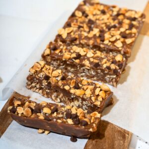 Delicious slices of no-bake peanut butter bars arranged on parchment paper atop a wooden chopping board, showcasing a rich, creamy texture and topped with chocolate chips and chopped peanuts.