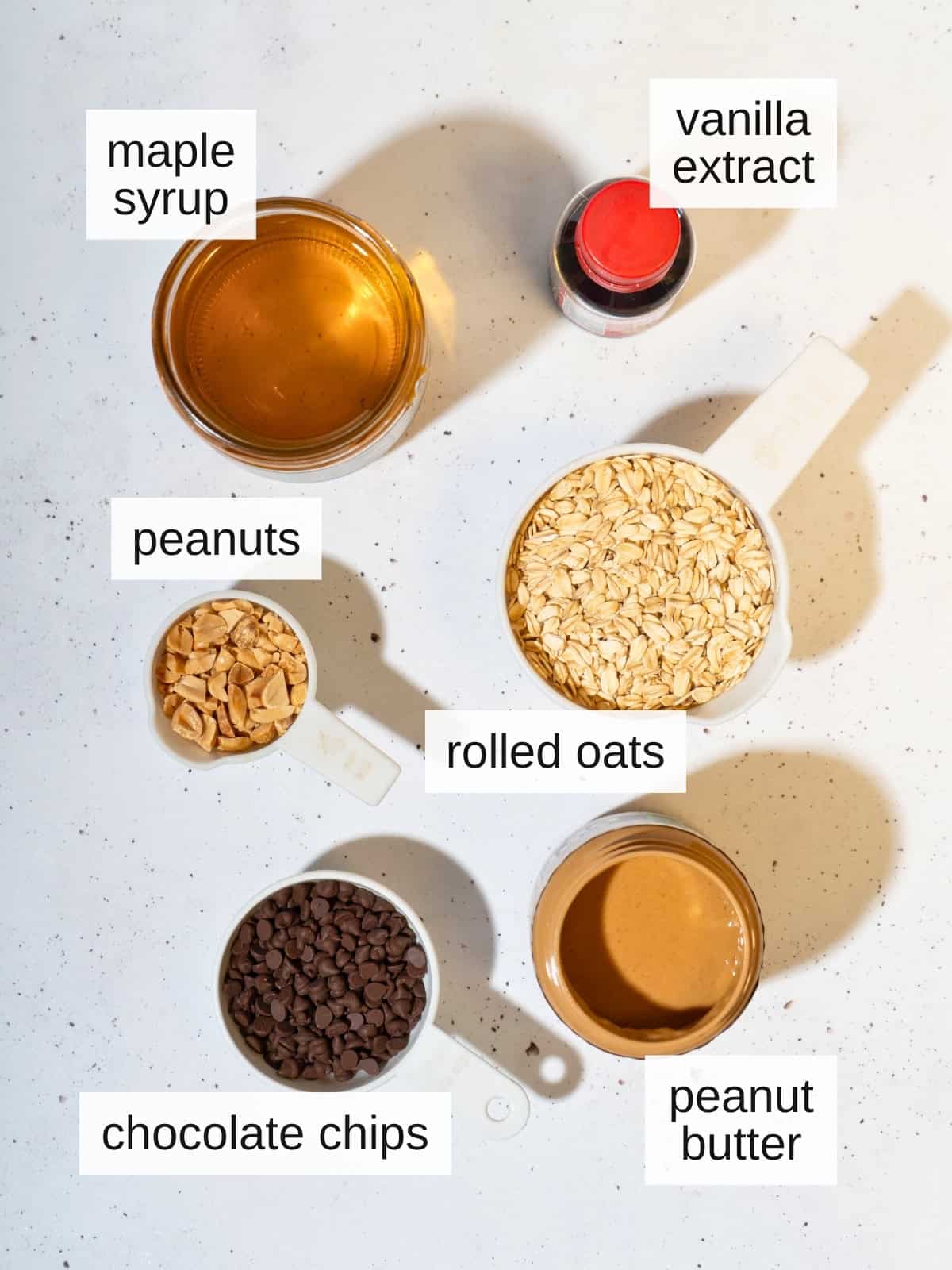 Ingredients used for peanut butter bars, including maple syrup, vanilla extract, peanuts, rolled oats, chocolate chips, and peanut butter.
